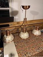 Full view of the chandelier showing three capiz shell mosaic shades mounted on copper-finished curved arms, resting on a patterned textile.