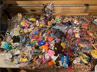 Photo showing a large assortment of mostly plastic McDonald's Happy Meal toys scattered on a table, some in original wrappers, various colors and characters visible.