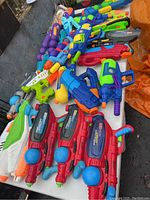 Photo of vintage Super Soaker water guns arranged on table showing multiple models including red XP 270 guns with blue spherical tanks, white and green water guns, purple multi-barrel gun.