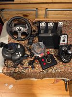 Photo showing Logitech G27 racing wheel, pedal set, gear shifter, Microsoft shifter handle, black box with joystick and buttons, various cables and USB adapters arranged on a patterned cloth surface.