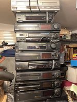 Stacked view of various VHS players from Sony, JVC, and Philips, showing front panels, control knobs, and brand markings.