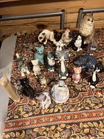 Photo showing nearly all 20 cat figurines arranged on a patterned fabric, including variety of porcelain cats, one black glossy cat, and a large beige cat resembling a Maine Coon.