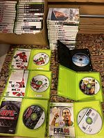 Photo showing stacked Xbox 360 game cases both in a large cardboard box and arranged outside it. Visible game titles include NHL 09, UFC 2009 Undisputed, FIFA 06, FIFA Soccer 08, Madden 2009 and others. Some cases are open showing discs and manual inserts inside.