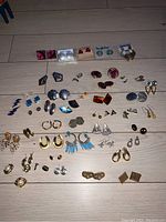 Photo showing multiple pairs of custom and vintage earrings arranged on a wooden surface, including studs, hoops, dangles in a range of colors and styles.
