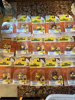 All 15 Minions figures shown in original packaging on patterned carpet background.