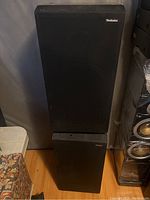 Photo showing front view of two stacked black Technics speakers in vintage style with fabric grills and visible wear on wooden sections.