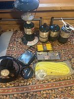 Vintage Coleman propane lantern with the damaged glass globe on top, three propane tanks, camping cook systems, fan, and other camping accessories arranged on a patterned fabric surface.