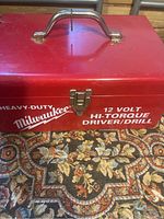Top view of red Milwaukee metal carrying case with silver handle and latch. Case marked with white text indicating 12 Volt Hi-Torque Driver/Drill.