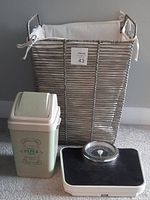 Photo of bathroom trash can, scales, and laundry basket grouped together on carpeted floor near gray wall.