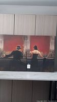 Two canvas paintings placed side by side showing a man and woman sitting at a bar with bottles behind them, against a red background.