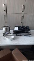 Photo showing the Trudeau raclette grill with stainless steel fondue pot, a white bowl, and base unit on a marble countertop.