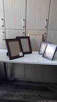 Photo shows five picture frames arranged on a white countertop against a light wood cabinet background. Two black picture frames of equal size (5" x 7") are placed next to each other, a larger black frame (7 1/2" x 9 1/2") stands behind them on the left, another black frame (10 1/2" x 8") is included, and a gold frame (13 5/8" x 10 5/8") is visible in the back.