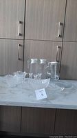 Glass serving items arranged on a countertop including large platter, small bowl, four candle holders, round serving plate, two pitchers, and one wine chiller.