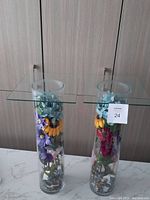 Two glass side tables with transparent square tops and cylindrical glass bases filled with colorful artificial flowers and decorative stones inside the bases, placed side by side against a wooden panel background.