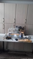 Full view of all kitchen items on a counter in front of cabinets, including utensils, glassware, bowls, and kitchen tools laid out and grouped.