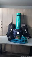 Photo showing all items on a kitchen countertop; two adult bicycle helmets (turquoise Giro and black Smith), blue sleeping bag, turquoise yoga mat, black camera bag, and a compact battery operated air pump.