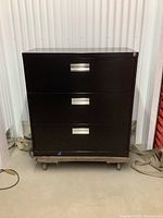 Front view of the black metal 3 drawer lateral filing cabinet on a dolly. Shows three horizontal drawers with handles and a key lock at top right.