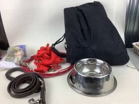 Photo showing black fabric dog car seat cover folded, multiple nylon harnesses and leads in red, brown leather leash, and stainless steel dog bowl.