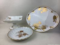 Three-piece lot of fine china dishes including one large oval plate with gold cornflower decorations, a square pedestal dish with gold floral accents, and an oval floral plate with gold trim.
