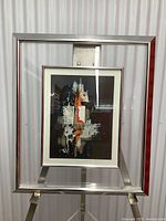 Frontal view showing the signed abstract art print in a clear glass frame with silver metal edges and stand.