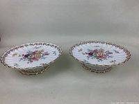 Two white oval pedestal serving dishes with multicolored floral Bouquet pattern, scalloped gold-edged rims, and gold-accented pedestal bases, placed side by side on a grey surface with neutral background.