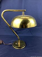 Side view of vintage brass desk lamp showing curved arm and round base with visible wear on brass surface.