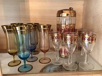 Full view of two sets of wine glasses in multiple colors alongside the pink and gold finished tall wine cooler or decanter on a mirrored shelf