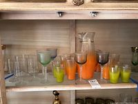 Wide view of entire glassware set on shelf showing large orange jug, various orange gradient glasses, smaller green glasses, clear glasses, and colored stem glasses.