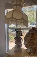 Full view of Capodimonte porcelain figurine lamp with cream fabric fringe shade in front of window.