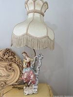 Full view of the Capodimonte lamp showing the porcelain figurine base and decorative lampshade with fringe.
