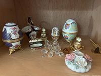 Shelf display showing hand-painted porcelain egg boxes, clear glass animal figurines, small decorative gold box, and porcelain flower piece.