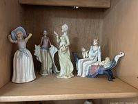Five porcelain figurines of women in various poses and attire on a wooden shelf.