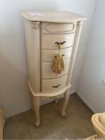 Front view of the cream-colored freestanding jewelry box with curved legs and tassel on middle drawer handle.