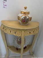 Photo showing the carved wooden half-moon entrance table in cream-beige color with floral carvings, the Limoges canister on top, and the decorative plate on lower shelf.