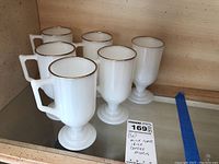 Six white opaque milk glass Irish coffee mugs with thick pedestal bases and squared handles, each rimmed with a thin band of 22K gold. Displayed on a shelf.