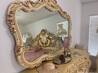 Full view of large ornate mirror on a wall above dresser, showing intricate carved frame with floral and scroll details.