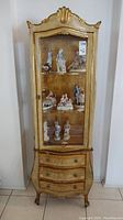 Full view of the tall carved wood Italian Florentine display cabinet with glass door showing interior shelves and figurines inside, includes curved base with three drawers.