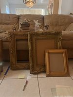 Four gold picture frames arranged side by side on floor in front of couch. Frames vary in size from small to large with ornate carvings or decorated edges
