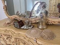 Wide shot showing dresser top with ornate mirror, crocheted hats, clock under glass dome, mirrored tray with perfume bottle and two candle holders