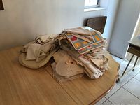 Pile of assorted table linens including various placemats, napkins, and tablecloths in neutral tones piled on a wooden table