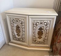 Front view of one cream and gold wooden bedside table showing carved decorative door panels with gold accents.
