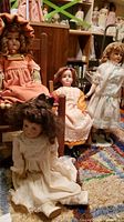 Four vintage dolls displayed in various dresses with visible features matching description; two wooden chairs partially visible in background.