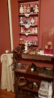 Four-shelf bookcase filled with dolls on upper shelves, assorted items on middle and bottom shelves, gowns hanging beside.