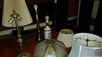 Four tall table lamps and various lamp shades arranged on a table and floor near a window
