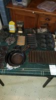 Overview of lot on green table showing cast iron skillets, kettle, flat iron, baking molds, trivets and tin match holders.