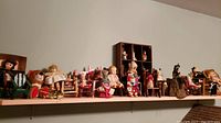 Full shelf with multiple dolls in different chairs including a vintage bisque doll, plush teddy bear, and dolls in wooden and painted chairs.