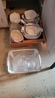 Multiple CorningWare dishes, lids, beige plastic strainer, and metal trivet seen in kitchen drawer and floor basket