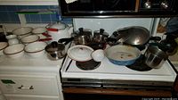 Photo of assorted enamelware pots and pans with red rims, Revere Ware stainless steel pots with lids, and a white enamel tea kettle.