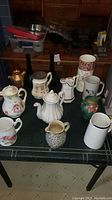 Overview of 10 ceramic pitchers and vases on green table, various designs and colors including floral and plain white with wear marks