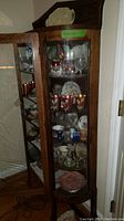 Full front angled view of the wood and glass curio cabinet showing curved door with glass and multiple display shelves filled with dishes and glassware (not included in sale).